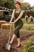 Woman in a green overalls holding a shovel in a field with another person in the background.